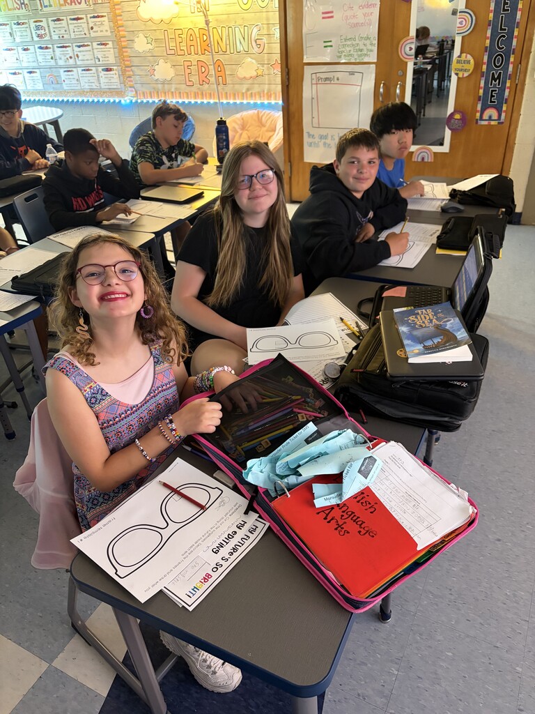 Four students sit in a row working on an assignment on paper while smiling for the camera