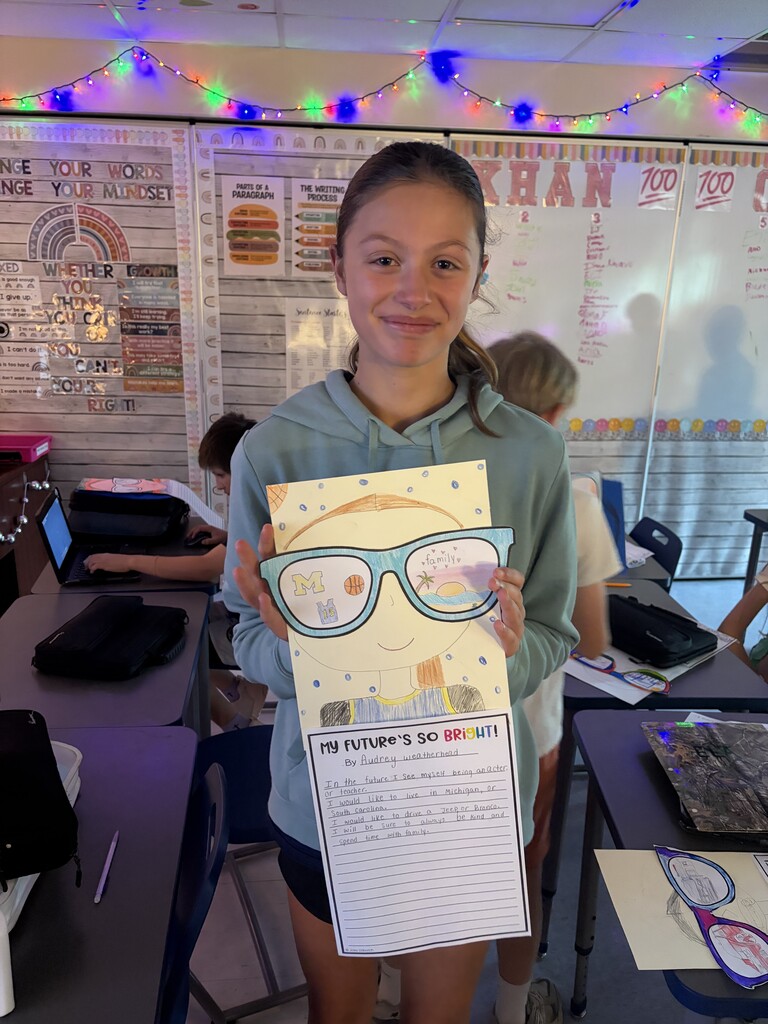 A student holds up her project with glasses on it and smiles for the camera in a classroom