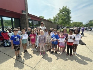 A large group of diverse, smiling kindergarten students standing outside on a sunny sidewalk with their female teacher. Several children are proudly holding up certificates or papers.