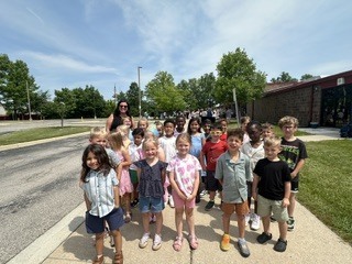 A large group of diverse kindergartens standing and smiling together on a sunny outdoor sidewalk, with the teacher standing behind them. They are positioned in front of a school building and parking lot under a bright blue sky.