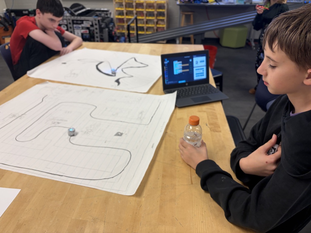 A student is siiting at a table looking at the drawing he made while holding a Gatorade bottle. There is a computer and another student in the background.