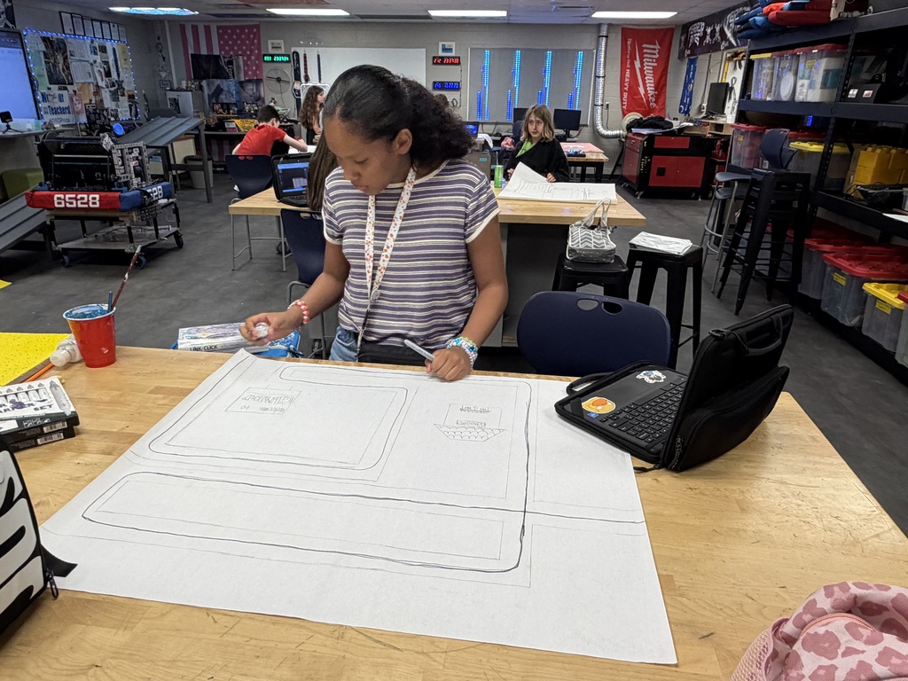 A student is standing at a table and working on her posterboard with her Chromebook. There are students working in the background in a STEM lab..