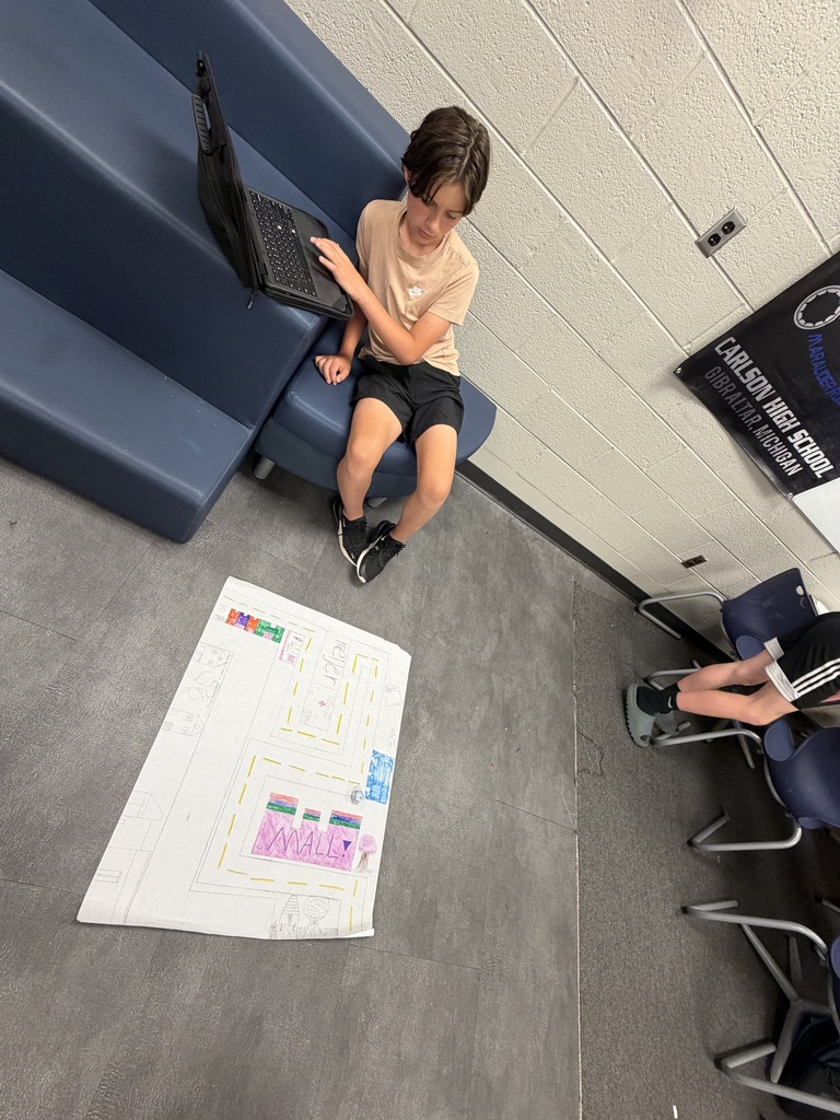 A student is sitting next to risers and working on his computer while looking at the posterboard he made which is sitting on the floor
