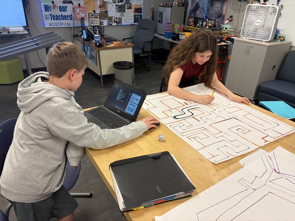Two students are sitting at a table. One is working on his computer and the other is drawing on her posterboard.