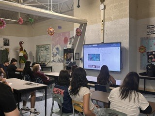 Students sitting at tables in a classroom watching a presentation on a smart board. 