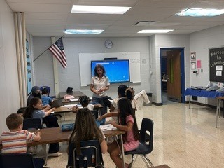 A teacher talking to elementary students as they are seated at tables in a classroom. 