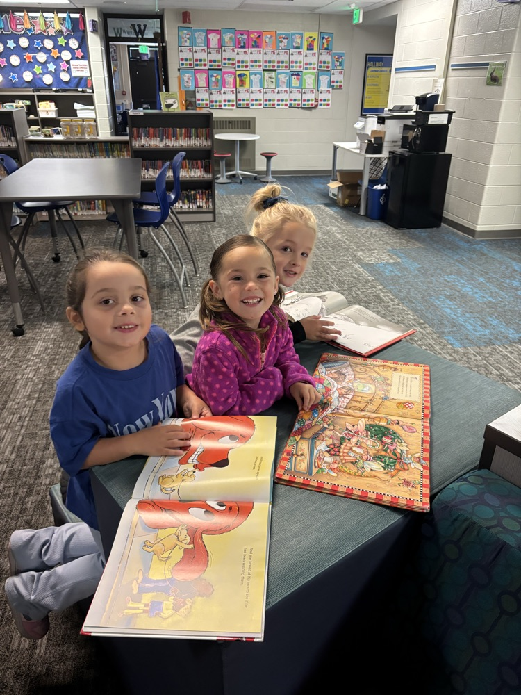 Kindergarten students from Ms. Brodeur’s class smile while holding and reading books during their first visit to the school library.