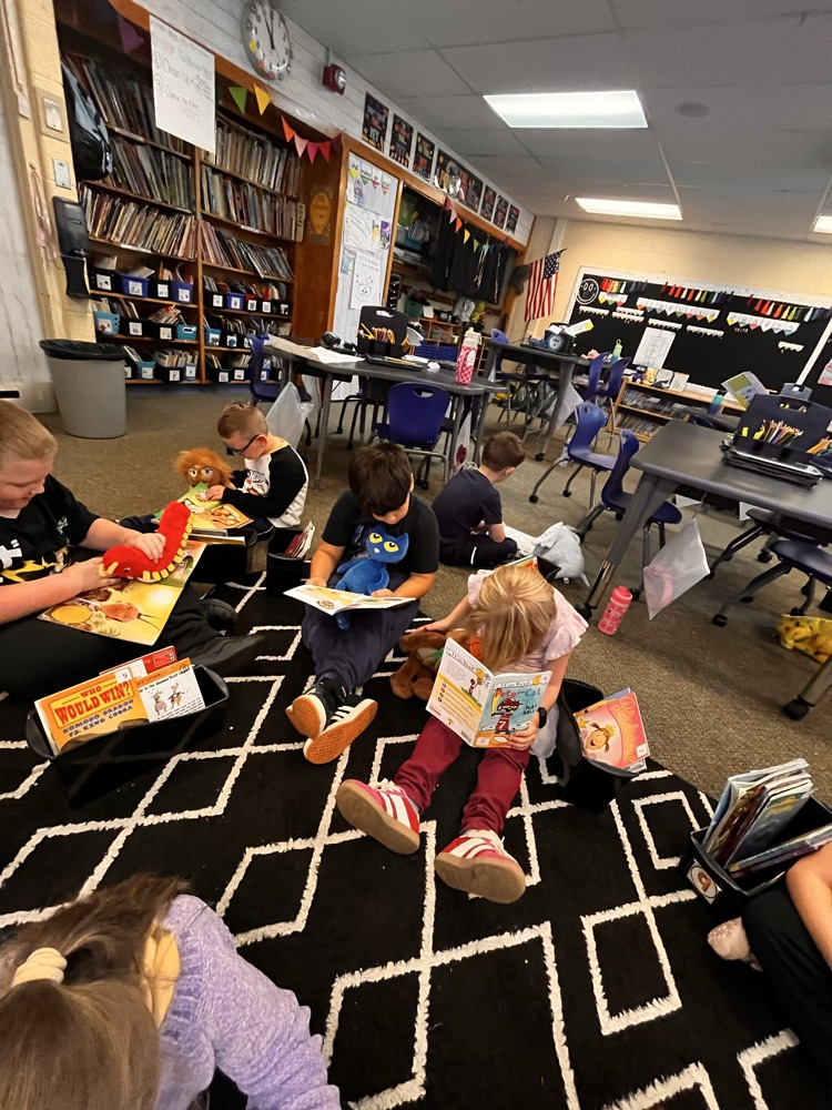 Photos show Mrs. Patrone’s 2nd grade students sitting in the classroom reading books while holding their stuffed animal buddies. Students are smiling and engaged with their books.