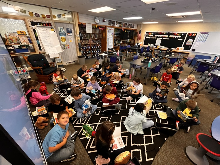 Photos show Mrs. Patrone’s 2nd grade students sitting in the classroom reading books while holding their stuffed animal buddies. Students are smiling and engaged with their books.
