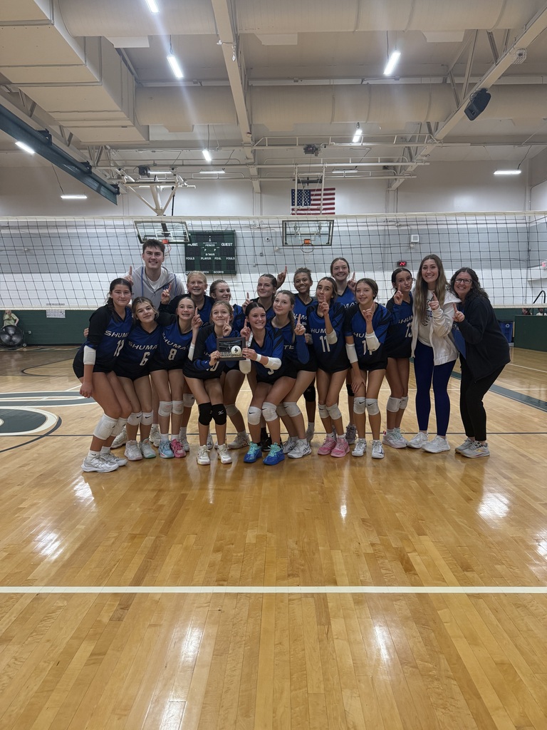 The 8th grade volleyball team stands in front of a volleyball net holding their trophy with their coaches
