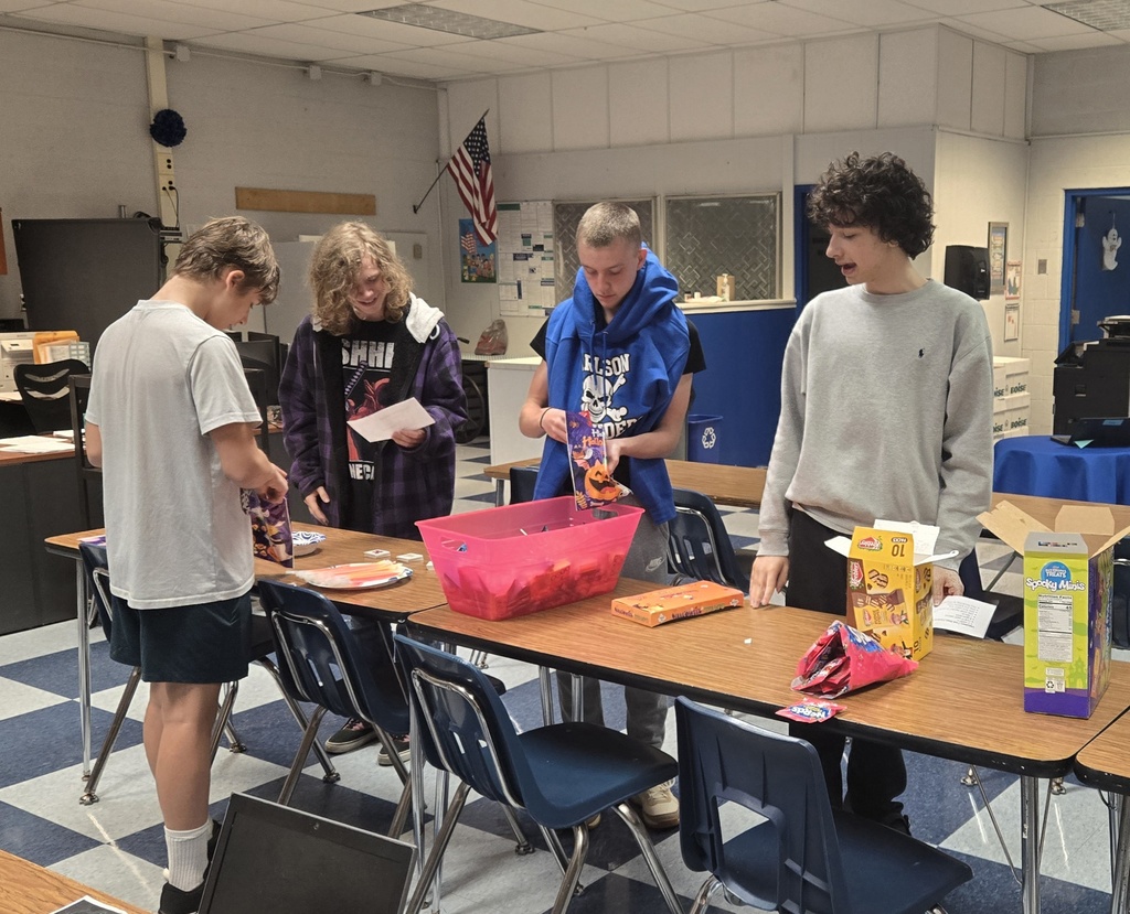 Four male students standing around a classroom table putting candy into Halloween bags. 