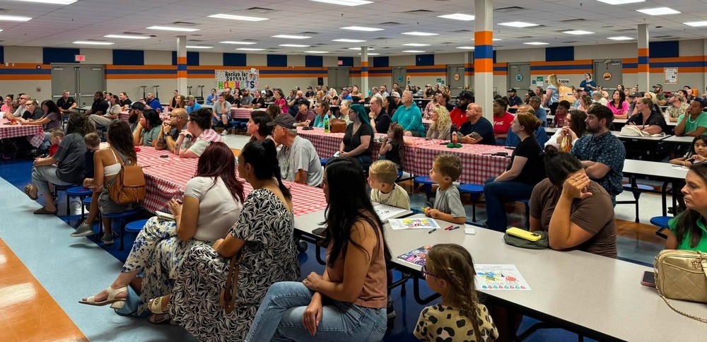 Parents, students, and community members gathered in a school cafeteria, seated at tables and benches, listening to a presentation during a large school event. Red-and-white checkered tablecloths cover several tables, and children are seated with adults throughout the room.