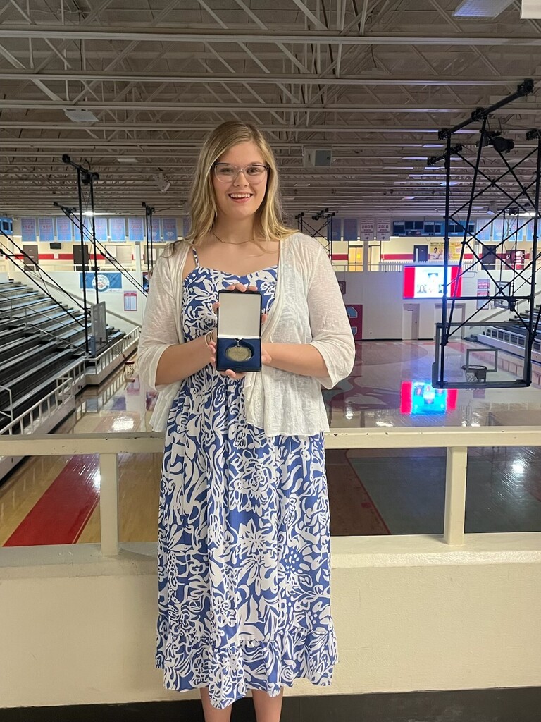 picture of a young, blonde girl dressed in a blue and white dress holding a medal in a case