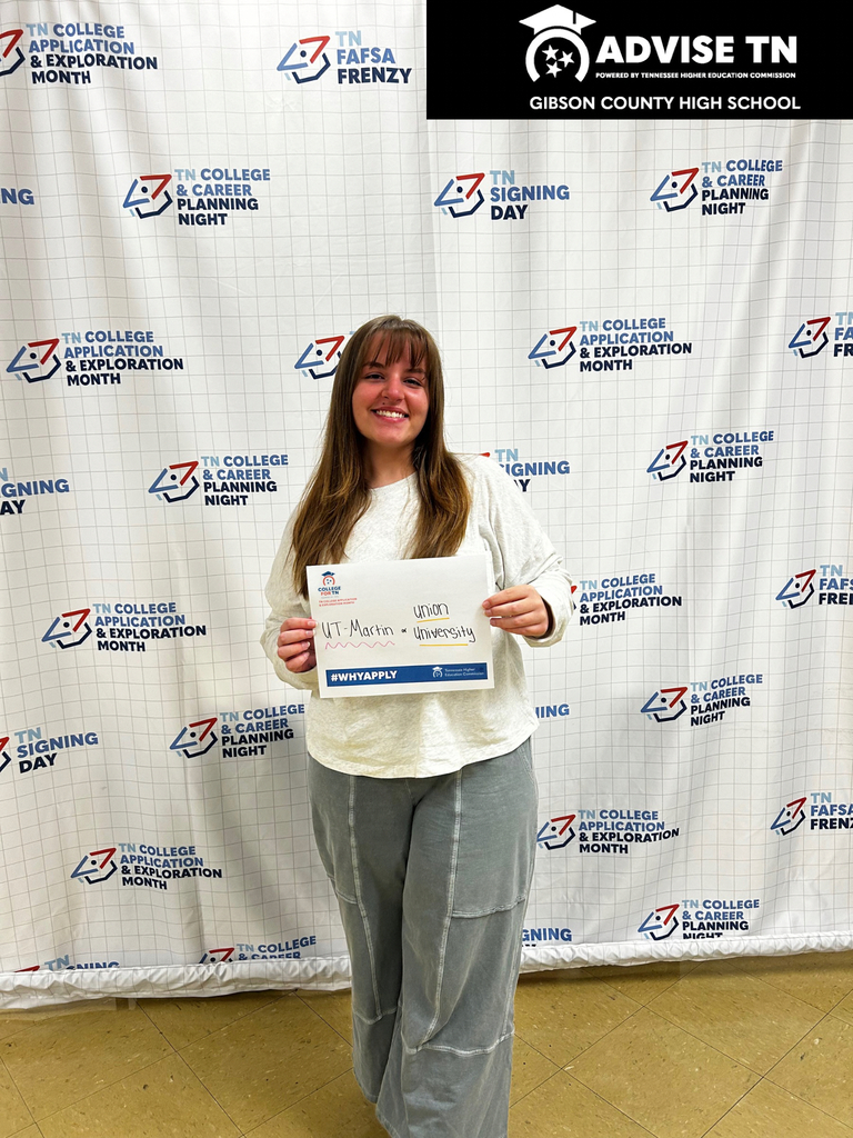 This image shows a smiling young woman standing centered in front of a white backdrop.   Subject: A young woman with long light brown hair and bangs is smiling directly at the camera. She is wearing a cream-colored crewneck sweatshirt and gray wide-leg cargo pants.   Holding Sign: She is holding a white paper sign with blue and red details. Handwritten in ink on the center of the sign are the names "UT - Martin" and "Union University". Below the handwritten text is a blue banner that reads "#WHYAPPLY".   Background: The backdrop features a repeating grid pattern of various educational initiative logos with blue and red icons. Text labels repeating across the banner include:   "TN College Application & Exploration Month"   "TN FAFSA Frenzy"   "TN College & Career Planning Night"   "TN Signing Day"   Top Header: Across the top right of the backdrop is a black horizontal banner featuring white text and a graduation cap logo that reads "ADVISE TN - POWERED BY TENNESSEE HIGHER EDUCATION COMMISSION" alongside "GIBSON COUNTY HIGH SCHOOL".   