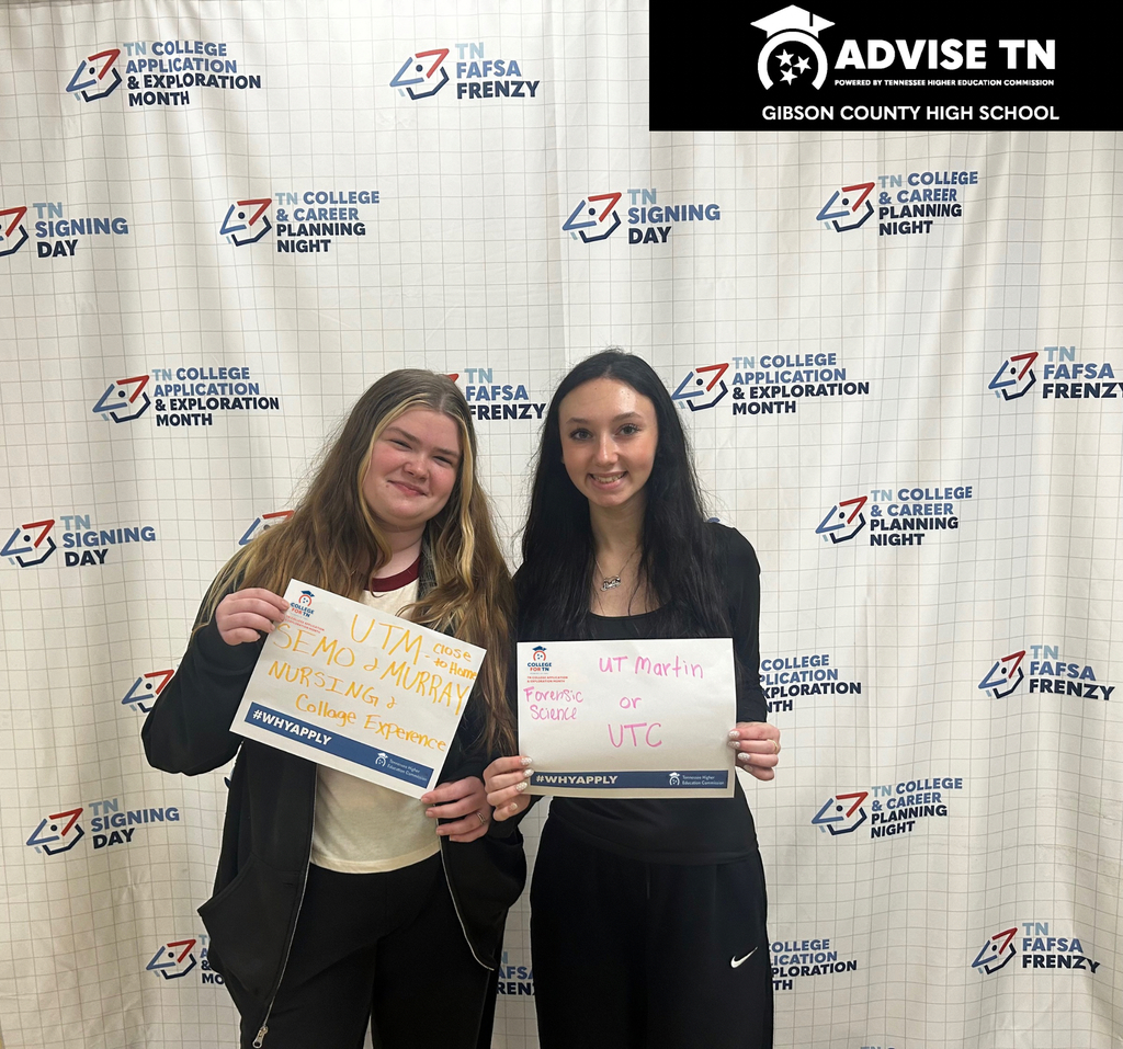 This image features two smiling young women standing side-by-side in front of an educational backdrop, each holding a small white sign outlining their college plans.   People & Signs (Left to Right):First Person: A young woman with long blonde hair wearing a black zip-up jacket over a burgundy shirt. Her sign has orange handwritten text that reads "UTM - close to home", "SEMO", "MURRAY", "NURSING", and "College Experience".   Second Person: A young woman with long dark brown hair wearing a black long-sleeve top. Her sign has pink handwritten text that reads "UT Martin or UTC", with "Forensic Science" noted on the left side.   Sign Details: Both paper signs feature a blue bar at the bottom with the hashtag "#WHYAPPLY" and Tennessee higher education logos.   Backdrop: The background is a white step-and-repeat banner patterned with blue and red logos for educational initiatives, including "TN College Application & Exploration Month", "TN FAFSA Frenzy", "TN College & Career Planning Night", and "TN Signing Day".   Top Header: Across the upper right corner is a black banner with white lettering that reads "ADVISE TN - POWERED BY TENNESSEE HIGHER EDUCATION COMMISSION" and "GIBSON COUNTY HIGH SCHOOL".   