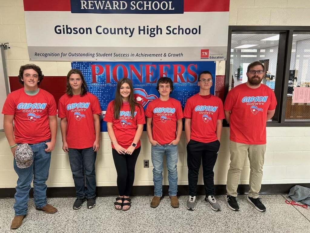Here is a descriptive summary of the image designed to be clear and accessible for someone who is visually impaired:Image SummaryOverviewThe image shows a group of six individuals—five young students and one adult supervisor—standing side by side in a hallway inside a school building. They are lined up horizontally in front of a light-colored brick wall with school signs displayed behind them.   Key Visual DetailsMatching Apparel:All six people are wearing matching bright red t-shirts printed with blue and white lettering that reads "GIBSON COUNTY" alongside a blue mascot graphic featuring a pioneer figure wearing a hat.   People (Left to Right):First Person (Far Left): A young male with short dark brown wavy hair, wearing blue jeans, brown cowboy boots, and holding a camouflage baseball cap in his right hand.   Second Person: A young male with shoulder-length light brown hair, wearing dark blue jeans and dark sneakers.   Third Person: A young female with long straight brown hair, wearing black leggings and open-toed strap sandals.   Fourth Person: A young male with medium-length brown hair, wearing light blue jeans and brown work boots.   Fifth Person: A young male wearing glasses with dark hair, black athletic pants, and gray and white sneakers.   Sixth Person (Far Right): An adult male with short brown hair, glasses, and a full beard, wearing beige khaki pants and dark sneakers with white soles.   Background & BannersMain School Banner (Top Center):A large white sign mounted on the wall framed by red bands at the top and bottom.   The top red band contains the text "REWARD SCHOOL" in white uppercase letters.   The center text reads "Gibson County High School" in large black font.   Below the name is the subtitle: "Recognition for Outstanding Student Success in Achievement & Growth" alongside the TN Department of Education logo.   Lower Banner (Behind the Group):A bright blue poster featuring a repeating pattern and large red letters spelling "PIONEERS".   Smaller white text printed across the middle displays key values/principles, including words like Integrity, Navigation, Empathy, Respect, and Service.   Environment:The setting is an indoor school hallway with light beige cinder block walls, a dark red trim stripe, and a speckle-patterned terrazzo floor. To the right is a interior window looking into an office space.   