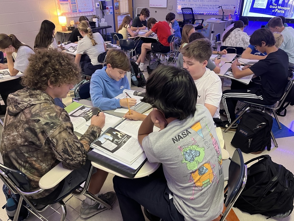 Middle school students work collaboratively in small groups at classroom desks, reading textbooks, discussing information, and completing written assignments about the Holocaust. Several students take notes while others examine reading materials. A classroom screen displays The Grand Mosque of Paris, connecting their group activities to a lesson on Holocaust history and literature.