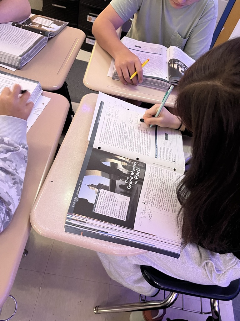 Middle school students work at their desks during a Holocaust literature lesson, reading and annotating informational texts. In the foreground, a student uses a pencil to make handwritten notes in a textbook featuring The Grand Mosque of Paris, while nearby classmates review passages and complete assignments. The activity emphasizes close reading, identifying textual evidence, and developing comprehension through written annotations.