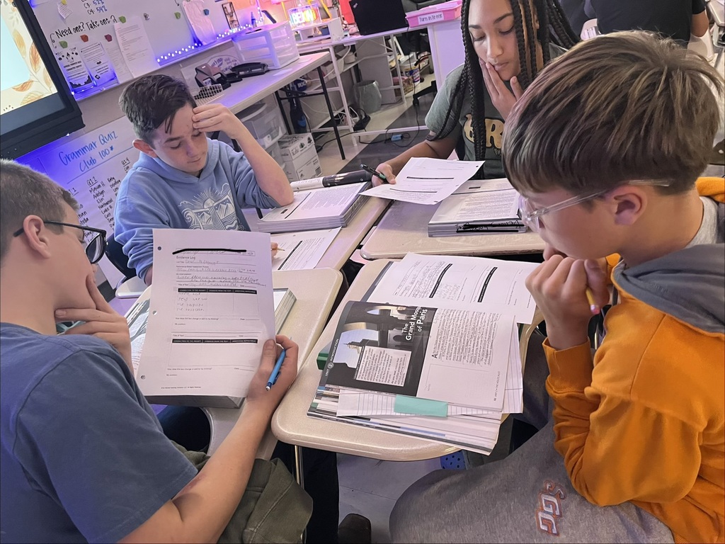 Four middle school students collaborate in a small group during a Holocaust literature lesson. Students examine an article titled The Grand Mosque of Paris, review evidence logs, and discuss written responses while seated at desks arranged together. Open textbooks, worksheets, and pencils are spread across the desks as students analyze informational texts and gather evidence to support their learning.