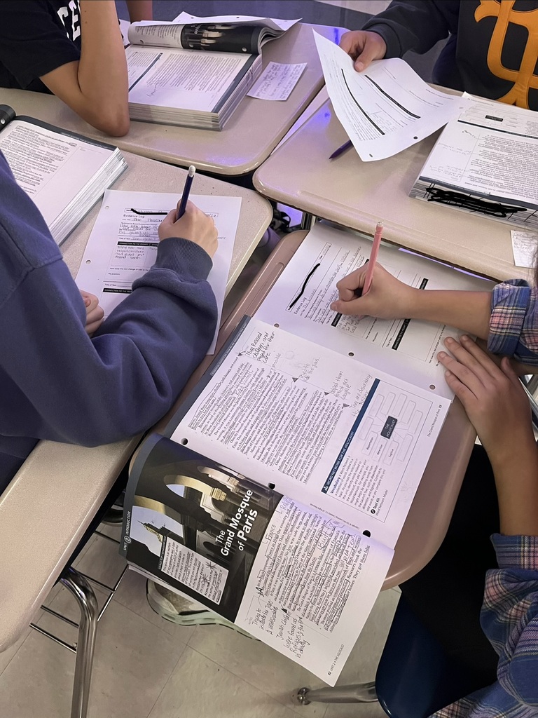 Middle school students work collaboratively at desks, reading and annotating an informational text titled The Grand Mosque of Paris. Students use pencils to complete evidence logs and record written responses while referencing passages in their textbooks. Open books, worksheets, and handwritten notes demonstrate students' engagement in close reading, text analysis, and gathering evidence during a Holocaust literature unit.
