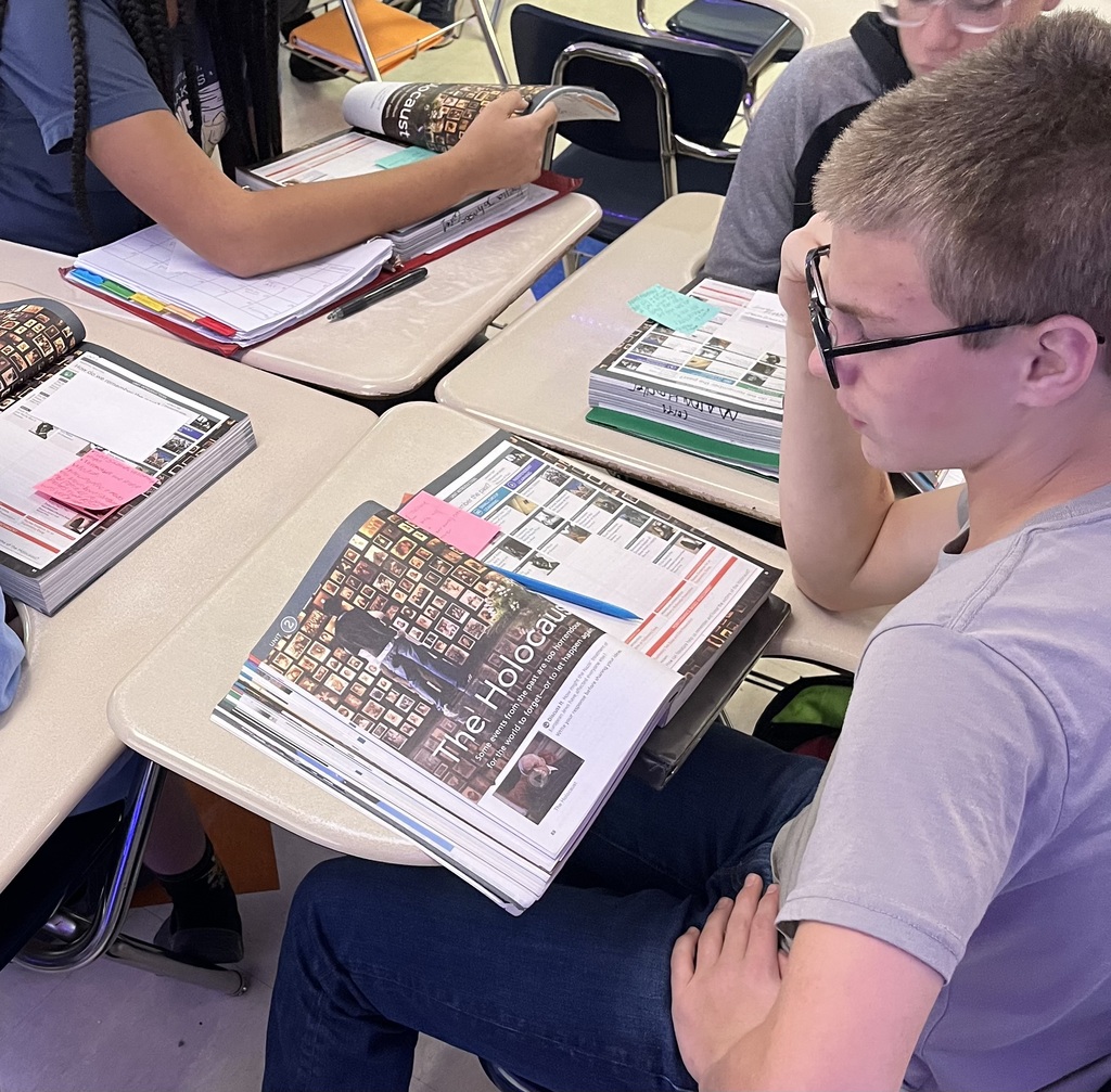 Middle school students sit at desks reading and examining textbook materials about the Holocaust. In the foreground, a student wearing glasses studies an open textbook featuring a Holocaust memorial photograph, while nearby classmates review similar materials. Colorful sticky notes, notebooks, and pencils are spread across the desks as students engage in a lesson about Holocaust history and remembrance.
