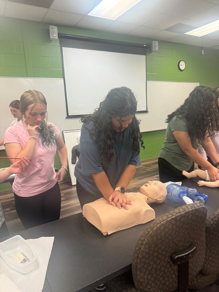 Middle school students participate in a hands-on CPR training activity. One student practices chest compressions on a CPR mannequin while others observe and practice nearby.