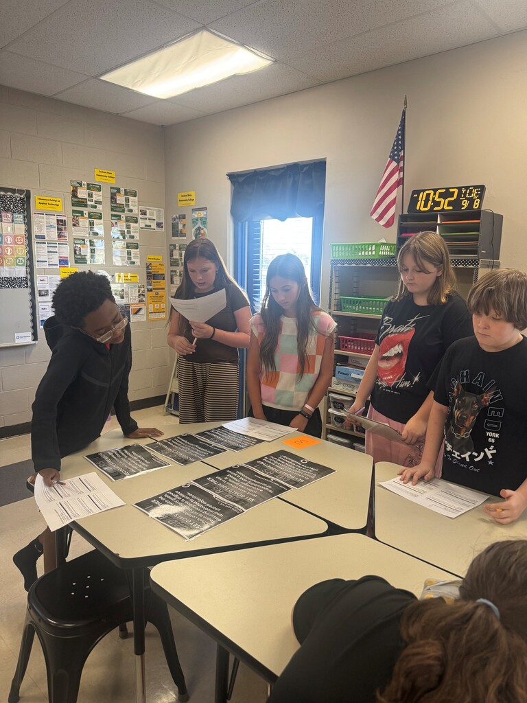 A group of middle school students stands around classroom tables, reviewing printed materials and discussing an activity together.