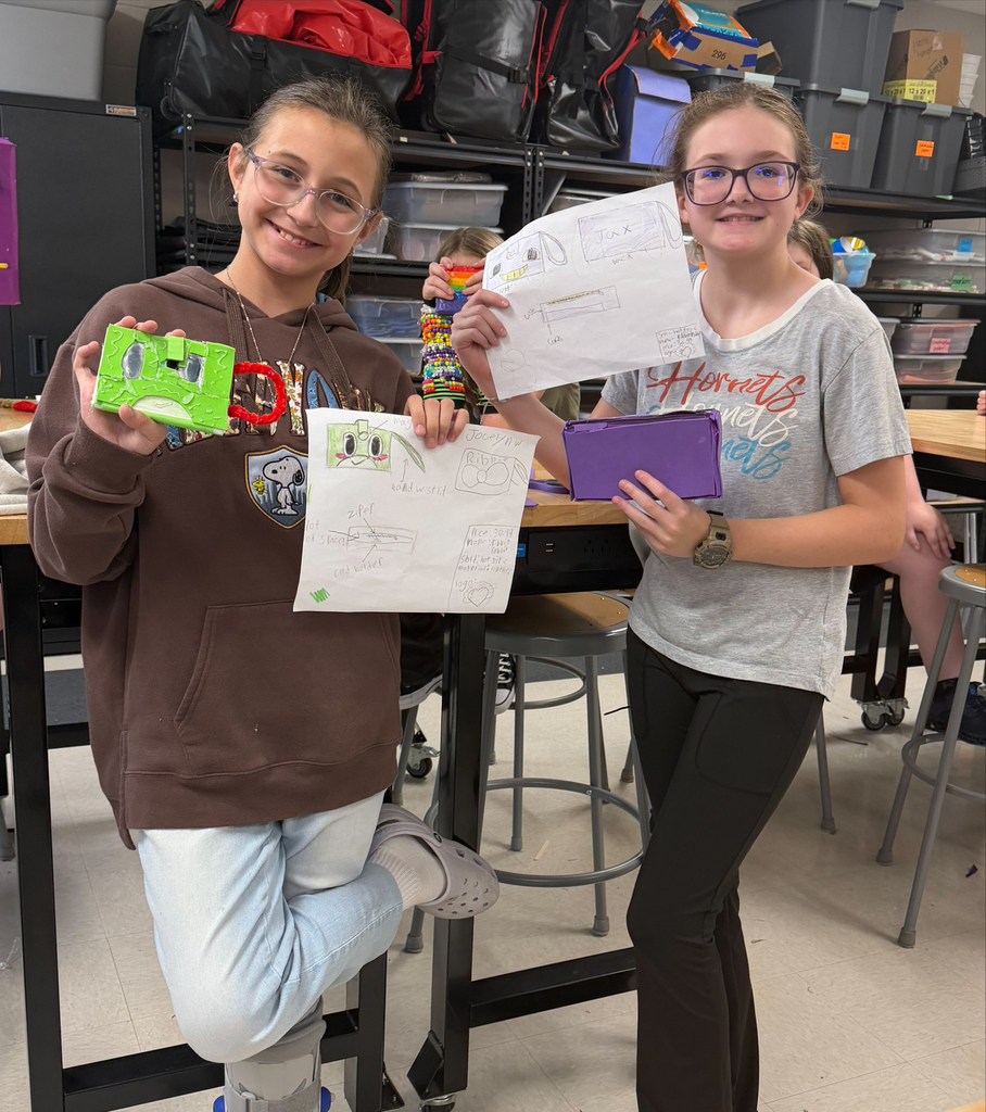Two middle school students smile while displaying their handmade prototypes and design sketches during a classroom engineering project.