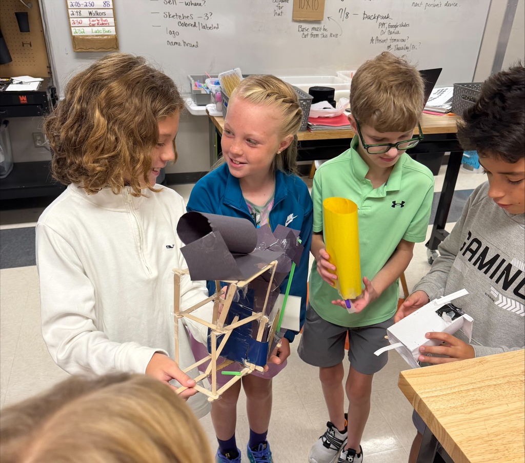 Four middle school students collaborate on a STEM engineering project, examining and sharing structures they built using craft sticks, paper, and other classroom materials.