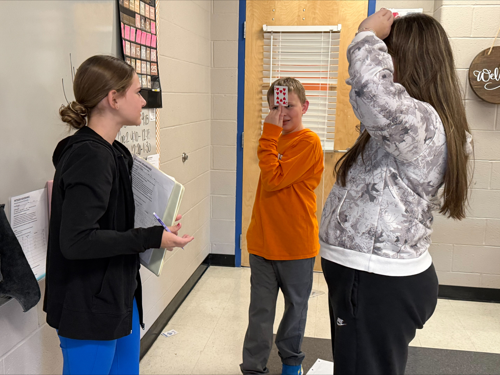 Three middle school students participate in a classroom card game near the classroom door. A student in an orange shirt holds a playing card against his forehead while two classmates face him, one holding a clipboard.