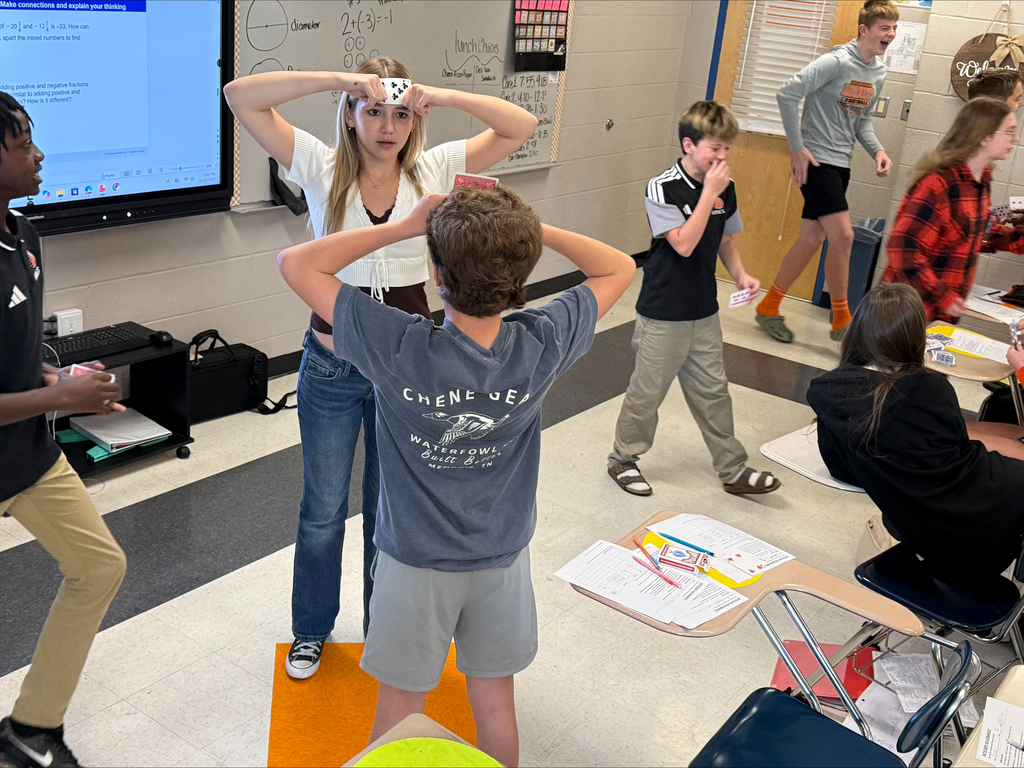 Middle school students engage in an interactive classroom card game. Two students face each other with playing cards held against their foreheads while classmates around them laugh, observe, and participate in the activity.
