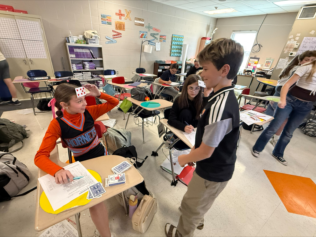 Middle school students participate in a collaborative classroom activity involving playing cards. A student in a cheerleading uniform holds a card against her forehead while a classmate stands nearby, with other students working at their desks.