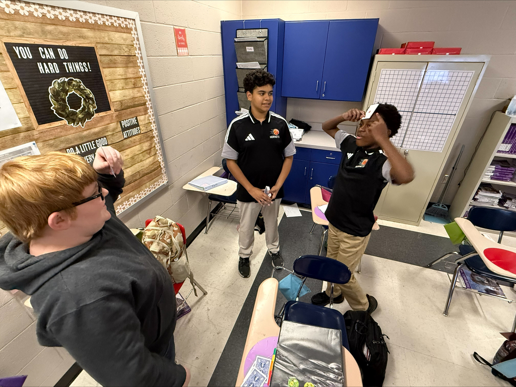 Three middle school students participate in an interactive card game in a classroom. Two students hold playing cards against their foreheads while a third student stands between them, observing the activity.