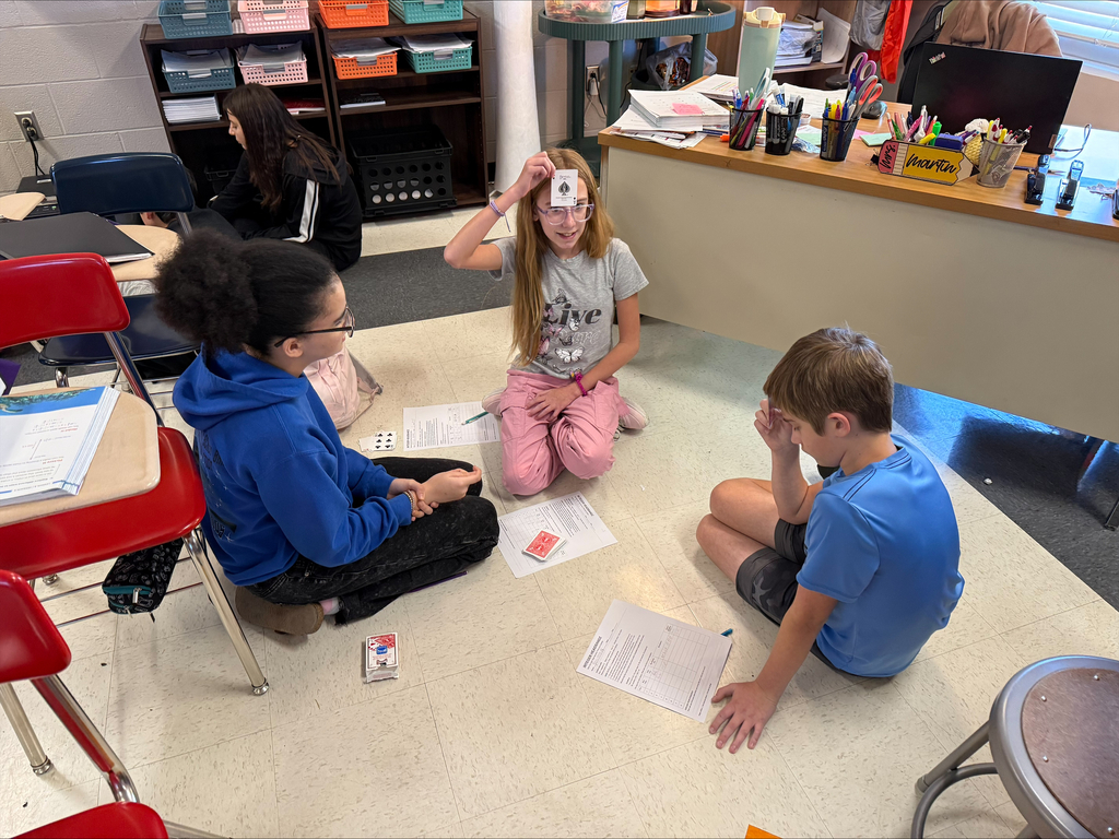Three middle school students sit in a circle on the classroom floor while participating in a playing card activity. One student holds a card against her forehead as the others observe, with worksheets and playing cards spread around them.