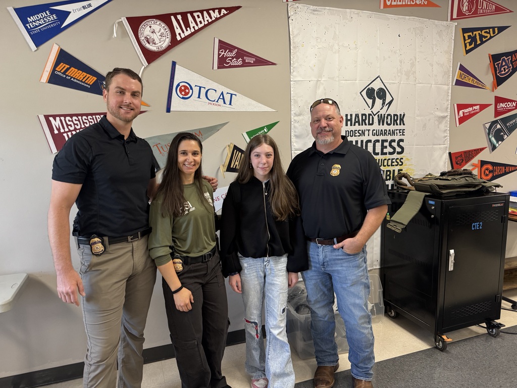 Three adults and a female student pose together in a classroom decorated with college and university pennants. Two adults wear badges on their belts, while the third wears a black polo with a badge emblem. A motivational poster about hard work and success hangs behind them.