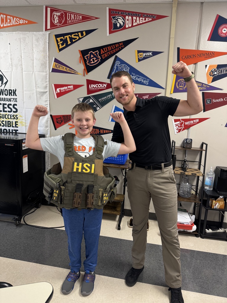 : A smiling male student wearing a tactical vest labeled “HSI” flexes his arms alongside an adult wearing a black polo, khaki pants, and a badge. The adult also flexes one arm. College and university pennants decorate the classroom wall behind them.
