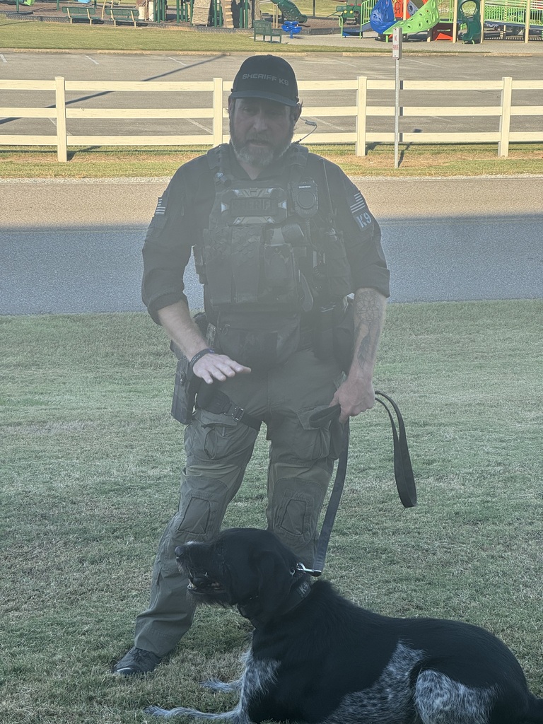A sheriff’s K-9 officer wearing a black tactical vest, cargo pants, and a cap stands on a grassy area while holding the leash of a large black-and-white dog lying in front of him. A white fence, road, and colorful playground equipment are visible in the background.