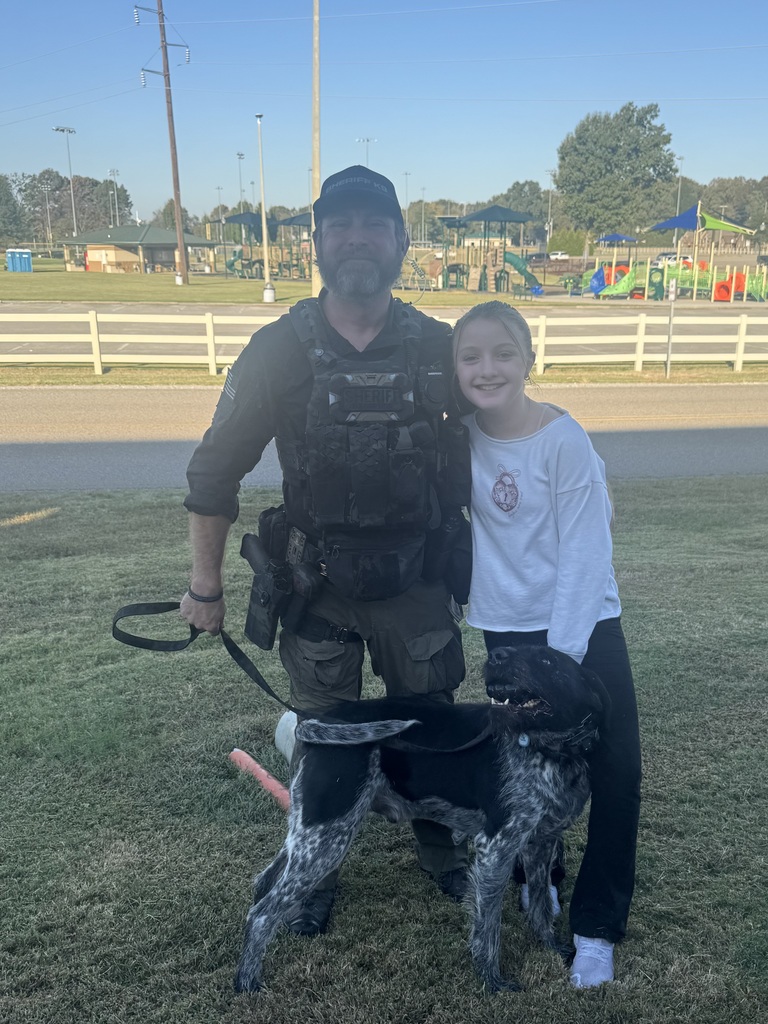 A smiling female student poses beside a sheriff’s K-9 officer and his black-and-white police dog outdoors. The officer wears tactical gear and holds the dog’s leash while the dog stands in front of them. A playground and white fence are visible in the background.