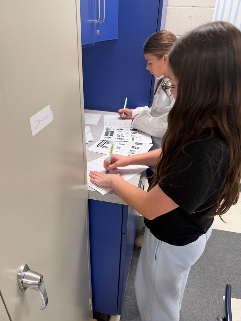 Two female students stand at a classroom counter completing worksheets for a math activity. They reference printed store advertisements while recording answers and calculations, with blue cabinets behind them.