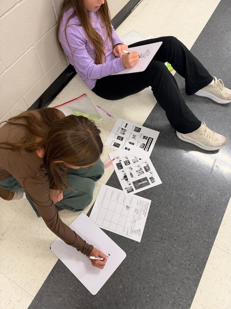 Two female students sit on the classroom floor using individual whiteboards to solve math problems. Printed store advertisements and worksheets are spread between them as they compare prices and complete calculations.