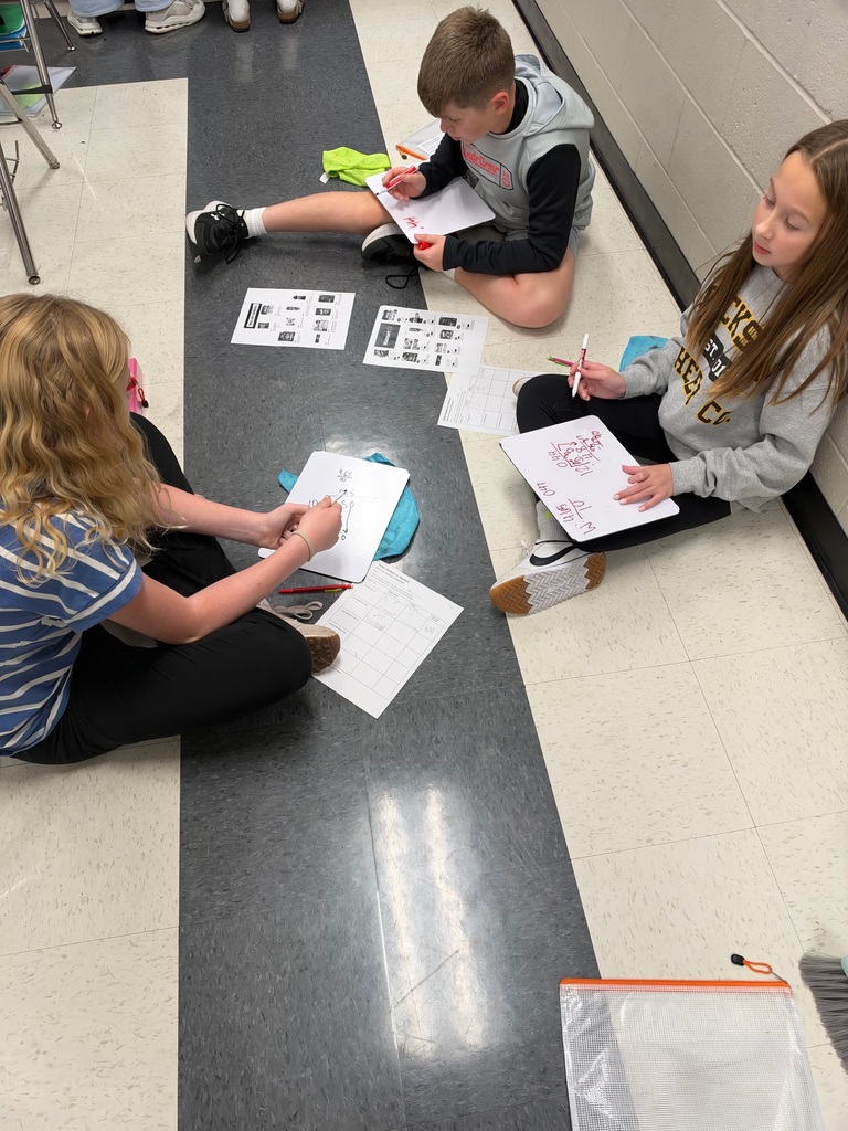 Three middle school students sit on the classroom floor working on a math activity. Each student uses a whiteboard to solve problems while referencing printed store advertisements and worksheets placed between them.