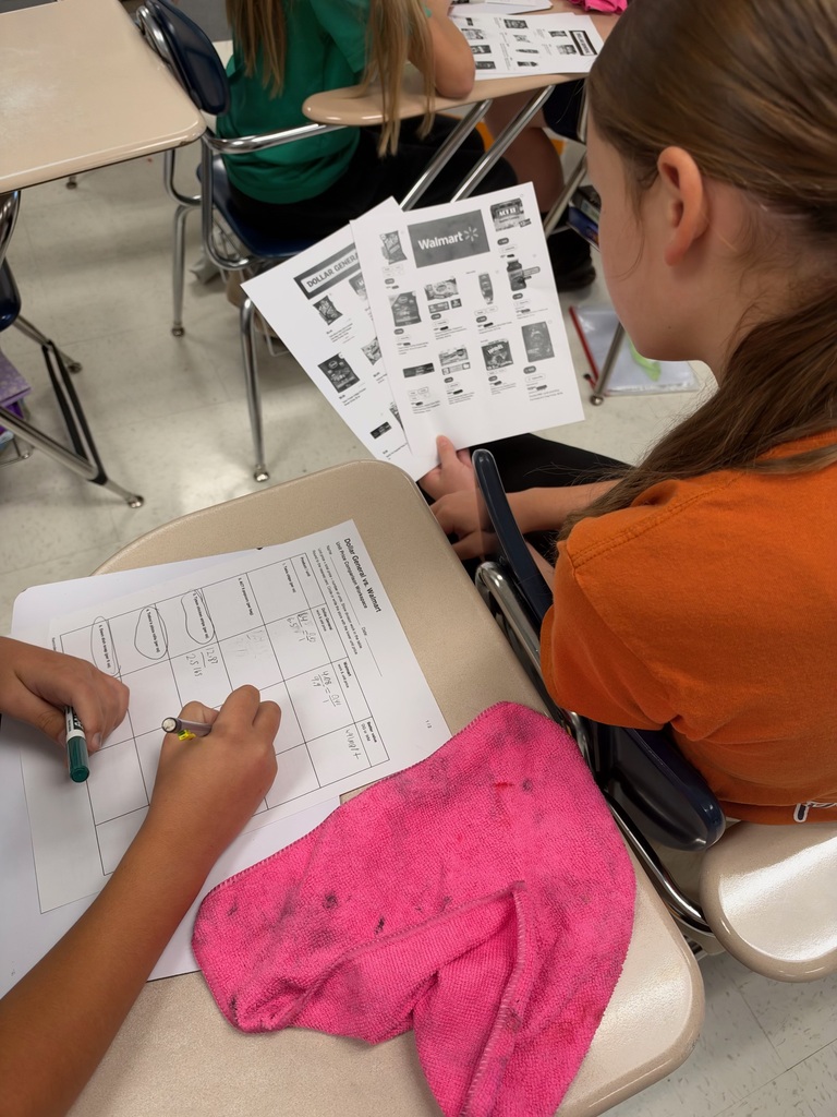 Two female students work on a math activity involving price comparisons between Walmart and Dollar General. One student examines printed advertisements while the other records calculations on a worksheet at her desk.