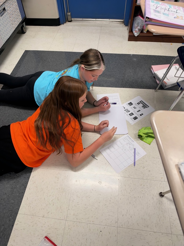 Two female students lie on the classroom floor while collaborating on a math assignment. They use a whiteboard, worksheets, and printed store advertisements to compare prices and solve problems.