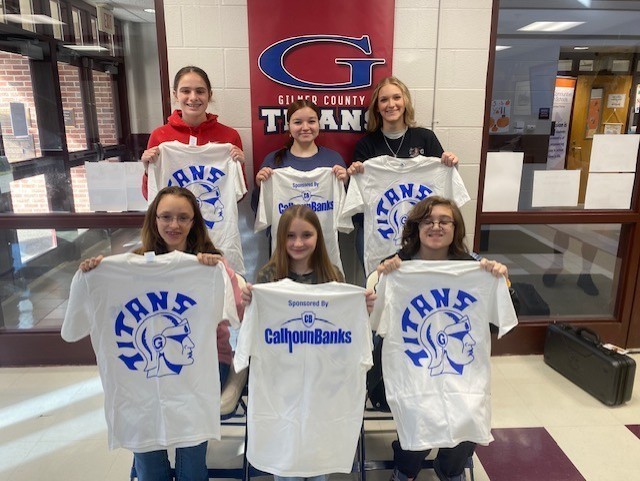 Six students standing indoors in front of a Gilmer County Titans banner, each holding white T-shirts featuring a blue Titan logo on the back and “Sponsored by Calhoun Banks” printed on the front.
