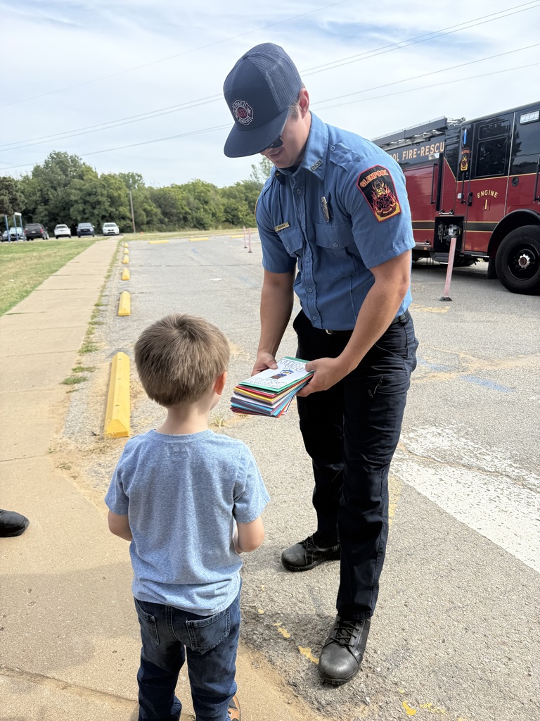 A firefighter hands books to a young boy on the side of the road. A fire engine is parked behind them.