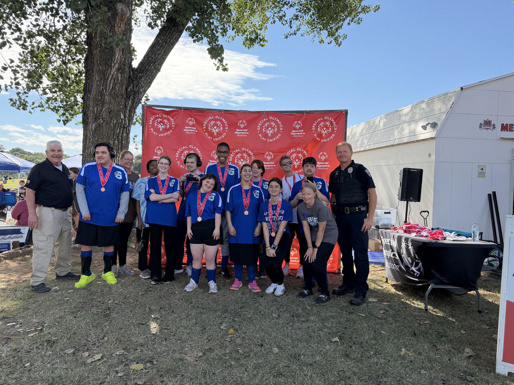 Group of students in blue jerseys and medals stand in front of a red banner. Behind them, a tree, a tent, and a speaker are visible.