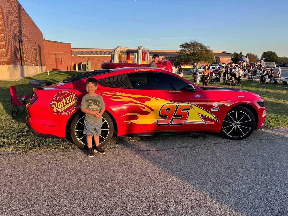 A boy poses beside a bright red, flame-covered Rust-eze race car with a large yellow 95, as families gather at a sunny school event.