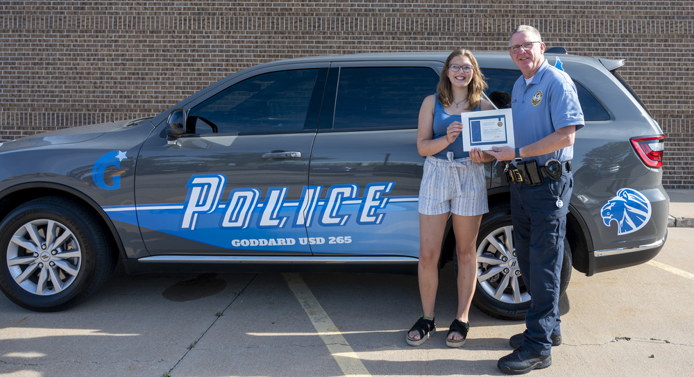 USD 265 Police Chief Joe Camp presents a certificate of appreciation to Taylor Harrington, recent Eisenhower High School graduate, on behalf of USD 265 for her design of the "car wrap" design that the USD 265 Police Department utilized to wrap all of their police vehicles - one of which is in the background behind Taylor and Chief Camp