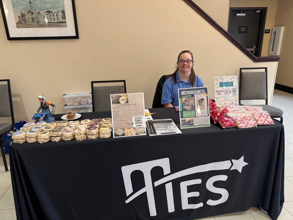 A woman sits behind a table with baked goods, flyers, and gifts displayed for sale. The tablecloth reads "TIES" with a star logo.
