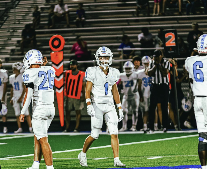 background: football field and stands with sideline players, players in white uniforms with blue numbers, EHS's Nathan Martens in center of photo